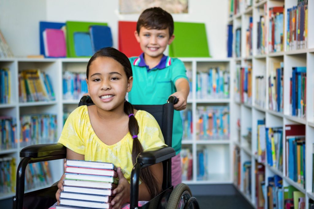 Uśmiechnięta dziewczynka trzyma w ręce kilka książek a za nią stoi również uśmiechnięty chłopiec